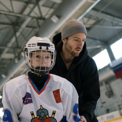 A father teaching his son to play ice hockey inside an arena, focusing on learning and bonding.