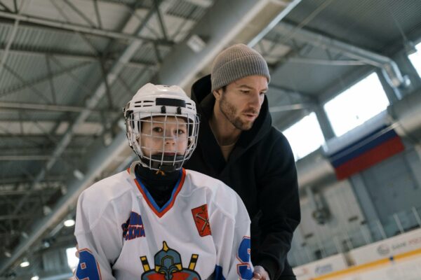 A father teaching his son to play ice hockey inside an arena, focusing on learning and bonding.